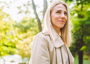 A representative photo of Lauren showing a person with long, straight blonde hair wearing a light beige trench coat, standing outdoors in a green, leafy park setting. Sunlight filters through the trees in the background, creating a bright and natural atmosphere.