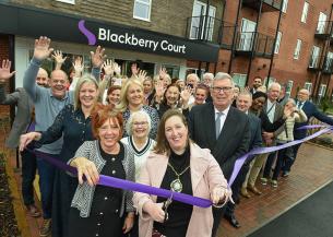 A group of people gathered outside a modern brick building with balconies, standing in front of a sign that reads “Blackberry Court.” They are participating in a ribbon-cutting ceremony, holding a long purple ribbon stretched across the entrance. Many individuals have their hands raised in celebration, and the setting appears to be the opening of a new facility or building.