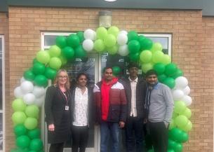 A group of five people stands in front of a Budgens store, which features a large arch made of green and white balloons. The individuals are smiling and dressed in casual clothing. The store's sign is prominently displayed above the entrance. The setting appears to be a festive occasion, likely a store opening or celebration.