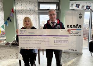 A man and a woman stand together in a bright, decorated room, holding a large novelty cheque made out to SSAFA - The Armed Forces Charity.
