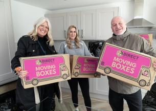 A group of three people smiling and standing in a modern kitchen, each holding a large cardboard box.