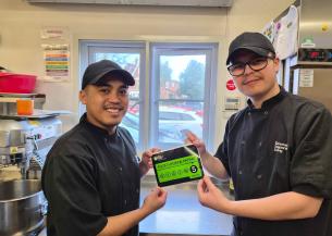 Two chefs in black uniforms and caps are standing in a kitchen. They are smiling and holding up a green food hygiene rating certificate that displays a score of 5. The kitchen is equipped with various cooking appliances and utensils, and there is a window in the background showing a view of parked cars outside.