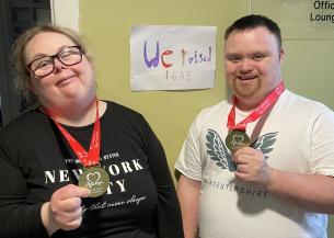 A woman and a man stand side by side, both smiling and holding medals. They are wearing red lanyards around their necks. Behind them is a sign that reads "We raised 1635," indicating a fundraising achievement.