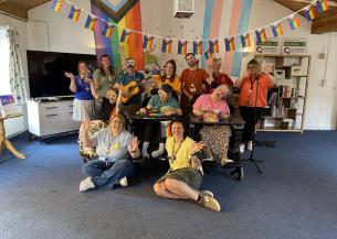 A group of people are gathered in a room decorated with colourful flags, including rainbow and trans pride flags.