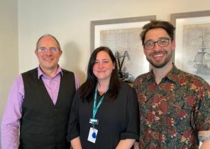 Three members of the Sanctuary Supported Living Learning and Development team standing indoors. One person wears a lilac shirt with a dark waistcoat, one wears a dark long‑sleeved top with a green lanyard, and one wears a patterned short‑sleeved shirt. They are standing side by side in front of framed artwork on a light‑coloured wall.