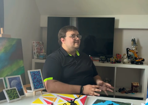 Person sitting at a desk with paintings and framed artwork, speaking in a home studio setting.