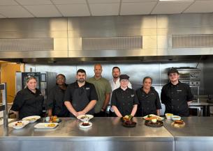 Catering colleagues stand behind a stainless-steel worktop displaying their completed dishes in a professional training kitchen.