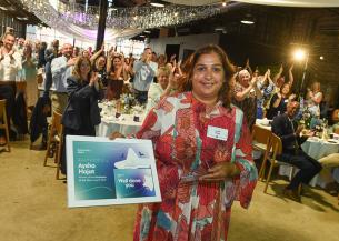 A woman in a red and white patterned dress stands smiling, holding a framed certificate. The certificate reads "Aysha Hajat, Employee of the Year." Behind her, a large group of people are standing and clapping in a decorated venue with draped fabric and lights. Tables are set with flowers and glasses, suggesting a celebratory event.