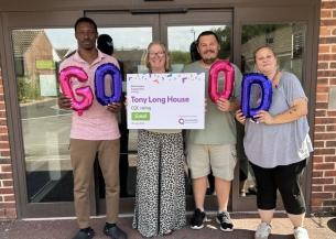 Four individuals stand in front of a building, holding balloons that spell "GOOD" and a sign with a "Good" CQC rating for Tony Long House.