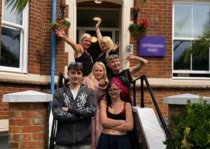 Group of six people standing on the steps outside a red brick residential building. Two people stand at the front with arms folded, while four others pose behind on the staircase. Hanging flower baskets frame the entrance with surrounding white-framed windows, black handrails and greenery.