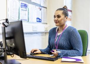 A young woman with a bun hairstyle sits at a desk, working on a computer. She is wearing a blue sweater and has a lanyard around her neck. The desk is organized with a computer monitor, a keyboard, a notepad, and a pen. Behind her, there are various documents and notices pinned to a board. The setting appears to be an office or study environment.