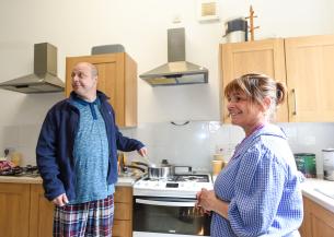 A man in a blue jacket and plaid pajama pants stands at a stove, stirring a pot. He looks to the side with a slight smile. A woman in a blue checkered shirt stands nearby, smiling back at him. The kitchen has wooden cabinets and modern appliances, with various kitchen items visible on the counter.