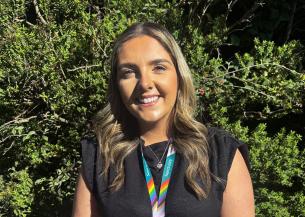 A young woman with long, wavy hair stands smiling in front of a lush green background. She is wearing a black top and a colorful lanyard with a rainbow design around her neck. The sunlight highlights her features, creating a warm and inviting atmosphere.
