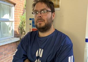 A man with glasses and a beard stands with his arms crossed, wearing a blue shirt with white stripes. A black geometric tattoo is visible on one forearm. He has a serious expression on his face. In the background, there is a poster on the wall that reads "CHANCES OF SUCCESS," and a window showing a glimpse of greenery outside.