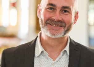 A smiling man with a beard is wearing a dark blazer over a light-colored, patterned shirt. He is standing in a well-lit indoor setting with soft, blurred background elements that suggest a warm and inviting atmosphere.