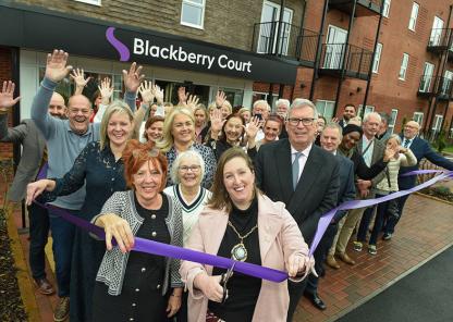 A group of people gathered outside a modern brick building with balconies, standing in front of a sign that reads “Blackberry Court.” They are participating in a ribbon-cutting ceremony, holding a long purple ribbon stretched across the entrance. Many individuals have their hands raised in celebration, and the setting appears to be the opening of a new facility or building.