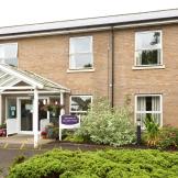 Two storey sandy brick-built retirement community located in Cambridgeshire. The entranceway has no step access. The building is decorated by a mixture of mature shrubbery and plants.