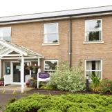Two storey sandy brick-built retirement community located in Cambridgeshire. The entranceway has no step access. The building is decorated by a mixture of mature shrubbery and plants.