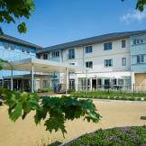 Contemporary, bright retirement living building. The bold blue pops of colour on a selection of the elevations gives a softer approach to the building against the white and grey. A small carpark features at the front of the property. 