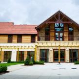 A luxurious property with a grand entrance and first storey balcony overlooking the white slabbed walkway. The property has been built using a mixture of natural materials such as brick, wooden panelling, and wooden beams. Small shrubbery plants line the perimeter.