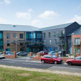 Located on a quiet corner plot the bold retirement living community façade consists of red, stone and grey brick and blue panelling on the larger exterior windows. Parking is located around the perimeter of the building.