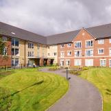 The newly built, yellow, and red bricked, three storey retirement living property set behind winding pathways which lead to a circular outdoor seating area as well the property and lovely grassed areas.