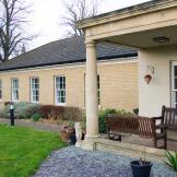 A period designed stone built building is home to the entrance of the property, the entrance is covered by a stone built porch with chunky columns supporting the roof. Two wooden benches sit underneath looking out onto the gardens. The rest of the building is built with soft coloured yellow bricks and long rectangular windows. 