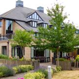 An idyllic brick built property with mass amounts of over sized windows and added skylights, set amongst a brick paved patio area and parasol seating enclosed in by wooden logged planters and colourful plants. 