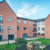 A modern red bricked new build retirement property. The building is built as a three storey corner plot with a small patio area along the left hand side of the property hosts an array of seating options from a wooden bench or metal chairs and circular tables. The patio leads into a circular tarmacked area with wooden planters and shrubbery, the rest of the garden is covered in grass.  