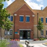 New build yellow brick built two storey property, with exaggerated triangular roofing, the entranceway is protruding the exterior designed in a red brick with mimicking pointed triangular roof. The outdoor area is planted with planted trees, flowers, and mature shrubbery.