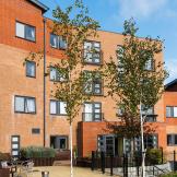 Beautiful contemporary apartment building contrasting dark grey window frames with the multitoned red bricked exterior. The outdoor area offers metal seating as well as numerous wooden benches all looking out to the newly planted trees and garden area.