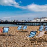 Striped wooden deck chairs placed in pairs on a pebbled beach facing out towards Brighton pier and the sea.