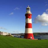 A bold red and white striped lighthouse placed on a grassy cliff, overlooking the Plymouth sound and city.