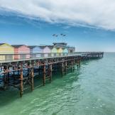 Multiple vibrant pastel and white striped beach huts lining either side of the Hastings pier, contrasting with the teal sea.