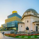 Stone built memorial hall with a small well kept and vibrant garden central to a large pedestrian walkway. In the background a four storey rectangular building with geometric pattern on the outside, alternating between a gold and blue glass colour scheme.