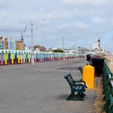 Concrete promenade walkway, to the left lined with a bold multi-coloured petite beach huts and to the right a shingled beach with multiple benches for a sea view.