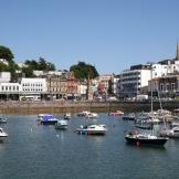 A busy harbour with numerous smaller boats decoratively displaying colourful buoys. A mix of properties cascade down from the hills towards the harbour.
