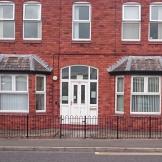 A red brick two storey building with bay windows on the ground floor set on a main road with a low black metal fence and gate in front of the building