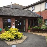 Multi elevational supported living property, built from red brick and a white rendered top elevation. A secure wooden leads into the property surrounded by mature shrubbery and large wooden planters