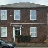 A detached, two storey brown brick built property, with a small brick built wall around the perimeter of the main building. Joined onto the building is a covered opening housing a parking space and access to the back of the property. 