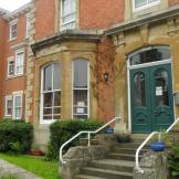 Exterior of a red brick terraced house with a bay window, a green double front door and steps leading down to a walkway