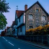 Stock image of a road with a lorry in the distance with a black and white tudor-style building next to a stone wall with metal railings and sunflowers poking over the top