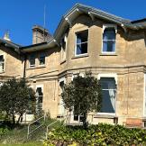 A large cream-grey building with two floors, with a green lawn in front and blue sky.