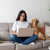 A woman sits on a plush grey sofa on her laptop with two labradors relaxing next to her