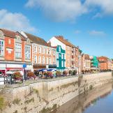 A row of colorful historic buildings along a riverside promenade on a bright sunny day, with reflections in the water