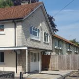 A two-story residential building with a light-colored exterior and a red-tiled roof is visible. In front of the building, there is a gated entrance leading to a parking area. The surrounding area features a stone wall and a wooden fence, with trees and greenery in the background. The sky is clear with a few clouds.