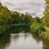 A calm river bordered by lush green trees and shrubs, with a church spire visible in the distance under a cloudy sky