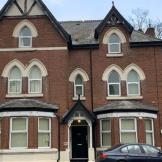 A large, three-story Victorian-style brick house with decorative gables and arched windows. The building features a symmetrical facade with a central entrance and two large bay windows on either side. The roof has a steep pitch, and there are trees in the background. A parked car is visible in front of the house, and the sky is partly cloudy.