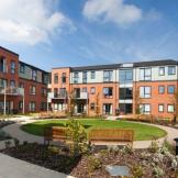 A modern, multi-story brick apartment building with large windows sits under a bright blue sky with scattered white clouds. A circular garden with a well-maintained green lawn and flowerbeds surrounds the building, featuring a wooden bench and pathway.
