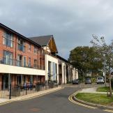 A street scene featuring a row of modern, multi-story brick apartments under a cloudy sky. A paved road curves gently ahead, with several parked cars and a yellow traffic cone visible. Trees and landscaping flank the road, and a small seating area is present at the front of the buildings.